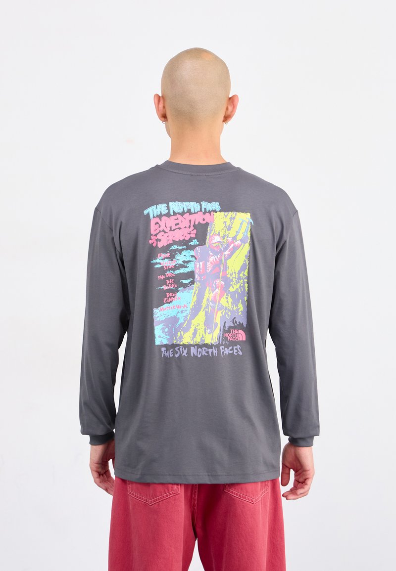 Bald person in gray long-sleeve shirt with colorful climbing design and text on back, wearing red pants, standing against white background.