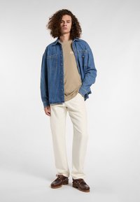 Denim shirt in blue with button closure and two chest pockets, beige t-shirt underneath, white trousers, and brown shoes with laces.