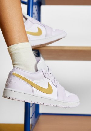 Personne portant des baskets Nike blanches et dorées et des chaussettes crème, assise les pieds posés sur des marches en bois avec une rampe bleue.