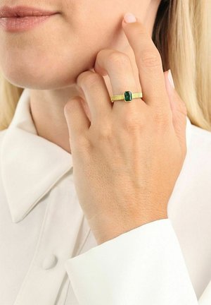 Donna che indossa un anello d'oro con una pietra verde rettangolare sul dito, toccandosi il viso, vestita con una camicia bianca con colletto.