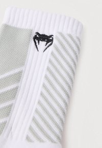 Biela a svetlozelená ponožka so striedavými pruhmi s textúrou, s čiernym logom kraba blízko vrchu. Pletená tkanina, priedušný dizajn.