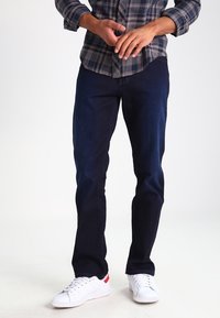 Mörkblå denimjeans med smal passform. Materialet verkar mjukt och slätt. Matchas med vita sneakers med röda detaljer.