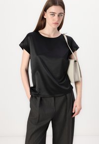 Zwarte satijnen top met korte mouwen en een ronde halslijn, gecombineerd met charcoal grijs gestreepte broek. Model houdt een lichtbeige tas vast.