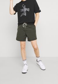 Černé tričko s šedým grafickým designem, olivově zelené šortky s tkaničkou a bílé tenisky s černými akcenty a pruhovanými ponožkami.