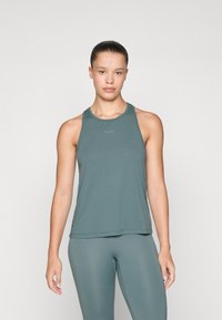 Vrouw met haar vastgebonden, gekleed in een teal kleurige mouwloze sporttop en bijpassende leggings, staande tegen een effen lichte achtergrond.