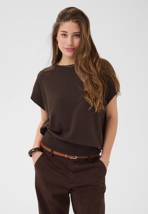 Jonge vrouw met lang golvend haar, gekleed in een donkerbruin top en broek met een gevlochten riem, handen in de zakken, zacht glimlachend tegen een lichte achtergrond.