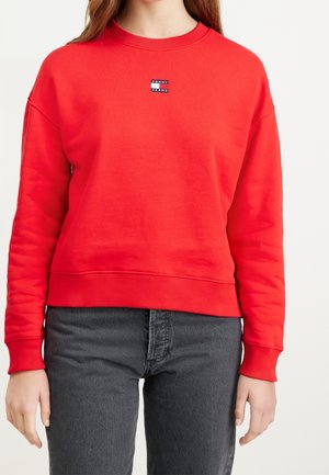 Kobieta w jaskrawo czerwonej bluzie Tommy Jeans z prążkowanymi mankietami i pasem, zestawiona z ciemnoszarymi dżinsami.