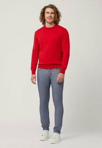 Maglione girocollo rosso in cotone abbinato a jeans skinny azzurri e sneaker bianche. Il modello con capelli ricci è in piedi contro uno sfondo neutro.