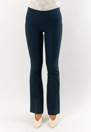 Donna che indossa pantaloni aderenti blu navy con una leggera svasatura abbinati a décolleté bianche con punta, in piedi davanti a uno sfondo neutro.