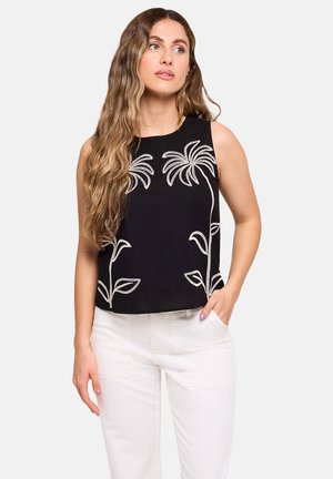 Mujer con cabello largo y ondulado que lleva una blusa negra sin mangas con bordado floral blanco y pantalones blancos, posando con una mano en el bolsillo.