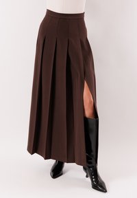 Gonna plissettata marrone scuro con spacco laterale che lascia intravedere la gamba, abbinata a stivali neri lucidi al ginocchio con punta affilata.