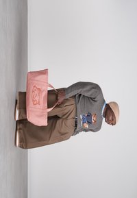 Ανδρας που φοράει μπεζ παντελόνι, γκρι πουλόβερ με σχέδιο αρκούδας, μπεζ σκούφο, κρατώντας μια μεγάλη ροζ τσάντα ώμου με εικονογράφηση αρκούδας.