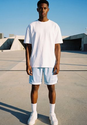 Junger Mann steht draußen und trägt ein weißes T-Shirt, hellblaue Shorts, weiße Socken und weiße Turnschuhe auf einer Betonfläche.