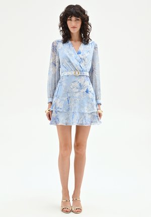 Donna con capelli ricci che indossa un abito a portafoglio azzurro chiaro con stampa floreale, cintura e maniche lunghe trasparenti, abbinato a sandali dorati con punta aperta.