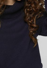 Cheveux bruns bouclés tombant sur un pull épais en maille bleu marine foncé avec un col montant, sur un fond clair uni.