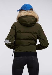Veste rembourrée vert olive avec capuche bordée de fourrure, poignets et bas côtelés noirs, et logo blanc sur la manche. Dispose d'un détail de fermeture éclair.