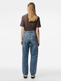 Sieviete ar gariem blondiem matiem, valkājot brūnu īsām piedurknēm apģērbu un vaļīgas, augsta vidukļa zilas džinsas, stāvot ar muguru pret skatītāju vienkāršā fona priekšā.