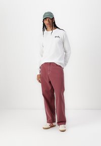 Long-sleeve white T-shirt with print, maroon baggy pants, and a green cap. Sneakers feature red stripes and a beige sole. Simple, modern design.