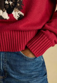 Červený pletený sveter s ribovaným lemom a manžetami; v kombinácii s džínsami, ktoré zobrazujú detail vrecka. Štruktúra vyzerá mäkká a teplá.