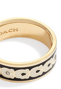 Bague plaquée or avec un bandeau noir ornée d'inscriptions en crème contrastant et d'une petite pierre claire. Fini lisse et poli.