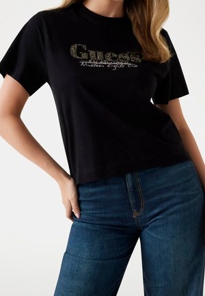 Kvinde iført en sort kortærmet top med "Guess Established Nineteen Eighty One" glitrende tekst og blå denimjeans.