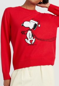 Rode gebreide trui met een cartoon van Snoopy in een kerstmanhoed en sjaal. Lange mouwen en geribde manchetten, gladde textuur.