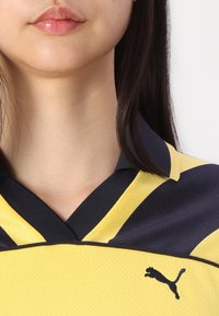 Gul och marinblå sporttröja med krage som har ett strukturerat mönster, svarta pipings och en Puma-logotyp på vänster sida.