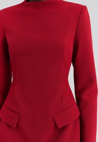 Piros, strukturált ruha hosszú ujjakkal, magas nyakkal és díszes, fodros zsebekkel a deréknál, texturált anyagból készült.