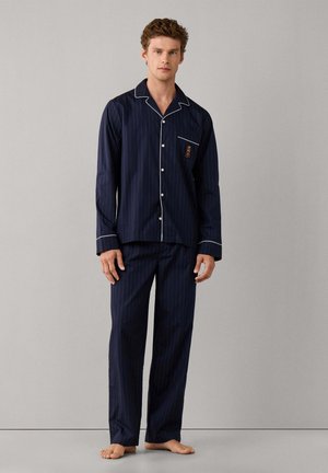 Jeune homme debout pieds nus portant un pyjama bleu marine à fines rayures, manches longues, avec un passepoil blanc et une petite broderie sur la poche poitrine.