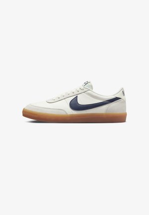 Zapatilla blanca de cuero con un swoosh azul marino, cordones planos, cuello acolchado y suela de goma. Diseño minimalista sin patrones visibles.