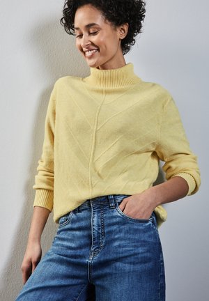 Gul strikket sweater med en mock turtleneck, der har skrå teksturmønstre. Parret med blå denim jeans, hænderne i lommerne.
