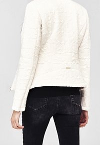 Hvid quilted jakke med præget skrift, der har en sort strikbånd. Synlige sømme og en sidelukning. Mørke jeans fuldender outfittet.