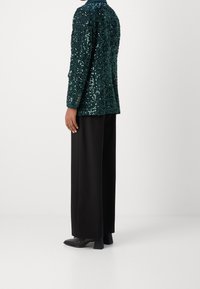 Teal paljettblazer med en slät finish, i kombination med svarta vida byxor och skor med blockklackar. Har knapppdetaljer vid ärmarna.