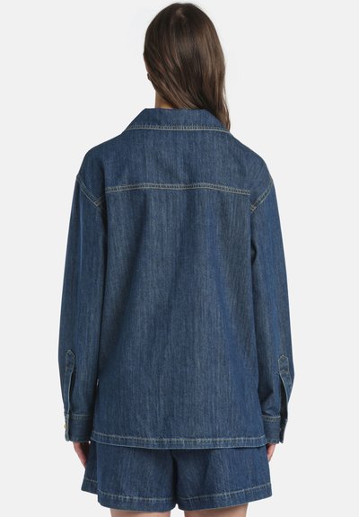 Personne aux longs cheveux bruns portant une veste en jean bleu oversize et un short en jean assorti, debout de dos face au spectateur.