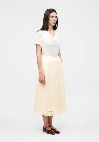 Γυναίκα που στέκεται φορώντας λευκή μπλούζα με κοντά μανίκια, κρεμ midi φούστα με δαντελωτό τελείωμα και σκούρα καφέ παπούτσια Mary Jane σε απλό φόντο.