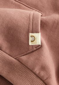 Hoodie w kolorze przydymionego różu, charakteryzujący się miękką fakturą materiału, prążkowanymi mankietami oraz wszywką z okrągłym wzorem w odcieniach brązu i kremu.