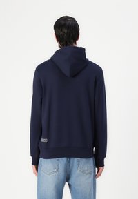 Felpa blu navy con cappuccio, polsini e orlo a costine. Presenta una patch con logo in vita. Il tessuto appare morbido e liscio.
