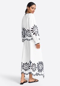 White dress set with black floral embroidery and scalloped hem. Long sleeves, relaxed fit, and a round neckline. Worn with flat sandals.