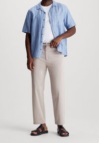 Chemise à manches courtes à rayures bleues portée sur un t-shirt blanc, associée à un pantalon beige et des sandales noires. Tissu lisse, coupe décontractée, design minimaliste.