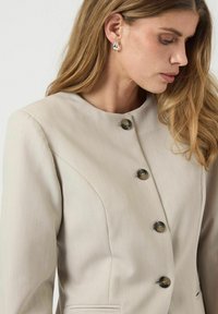 Beige blazer med rund krave, udstyret med fem sorte knapper, vandrette søm og sidelommer. Glat stof med en subtil glans.