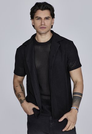 Veste noire à manches courtes et côtes avec un col, sur un haut noir rayé transparent. Le mannequin porte un jean foncé et arbore des tatouages.