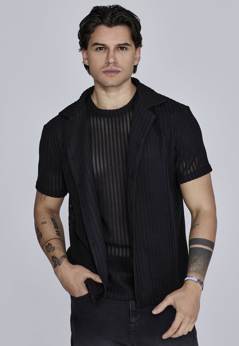 Chaqueta negra de canalé de manga corta con un diseño de cuello sobre una blusa negra de rayas transparente. La modelo lleva jeans oscuros y muestra tatuajes.