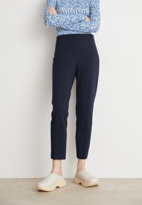 Person står med armarna i kors iklädd en blåmönstrad långärmad topp, marinblå byxor i ankellängd och beige slip-on skor med texturerade sulor.