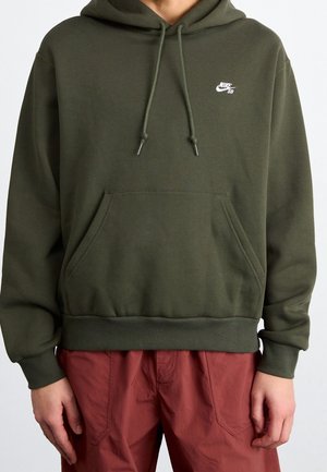 Personne portant un sweat à capuche Nike SB vert olive avec poche avant et cordons, associé à un pantalon rouge sur un fond uni.