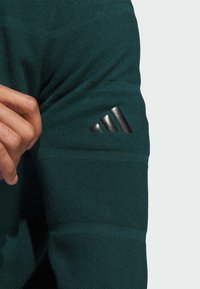 Mørkegrønn strikket langermet plagg med teksturert overflate. Har en svart, tre-stripe logo på overarmen.