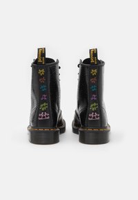Botas negras hasta el tobillo con una superficie texturada, presentando figuras de dibujos animados coloridas en la parte posterior y una lengüeta de logo amarillo en el talón.