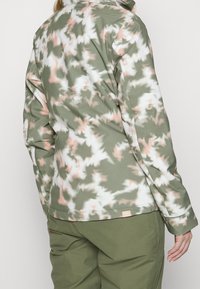 Grüne und weiße gemusterte Jacke mit weicher Textur, hoher Kragen und einem Logopatch am Saum. Kombiniert mit grünen Hosen.