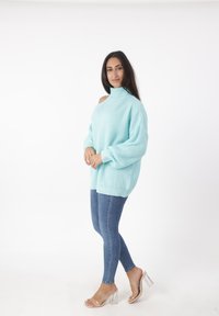 Donna in maglione azzurro chiaro a maglia con spalla scoperta, jeans skinny blu e sandali trasparenti con tacco su sfondo bianco.