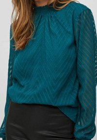 Blusa teal con un motivo a zigzag testurizzato, maniche lunghe trasparenti e scollatura arricciata, abbinata a una gonna nera di pelle.