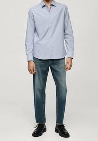 Mies, joka on pukeutunut vaaleansiniseen raitapaitaan, tummansinsisiin suoriin housuihin ja mustiin nahkakengissä, seisoo yksinkertaista taustaa vasten.